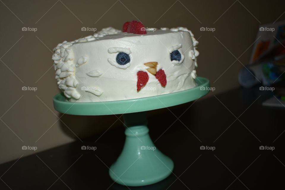 A feisty chicken cake with natural white icing and fruit for decoration sits atop a turquoise cake stand against a neutral background