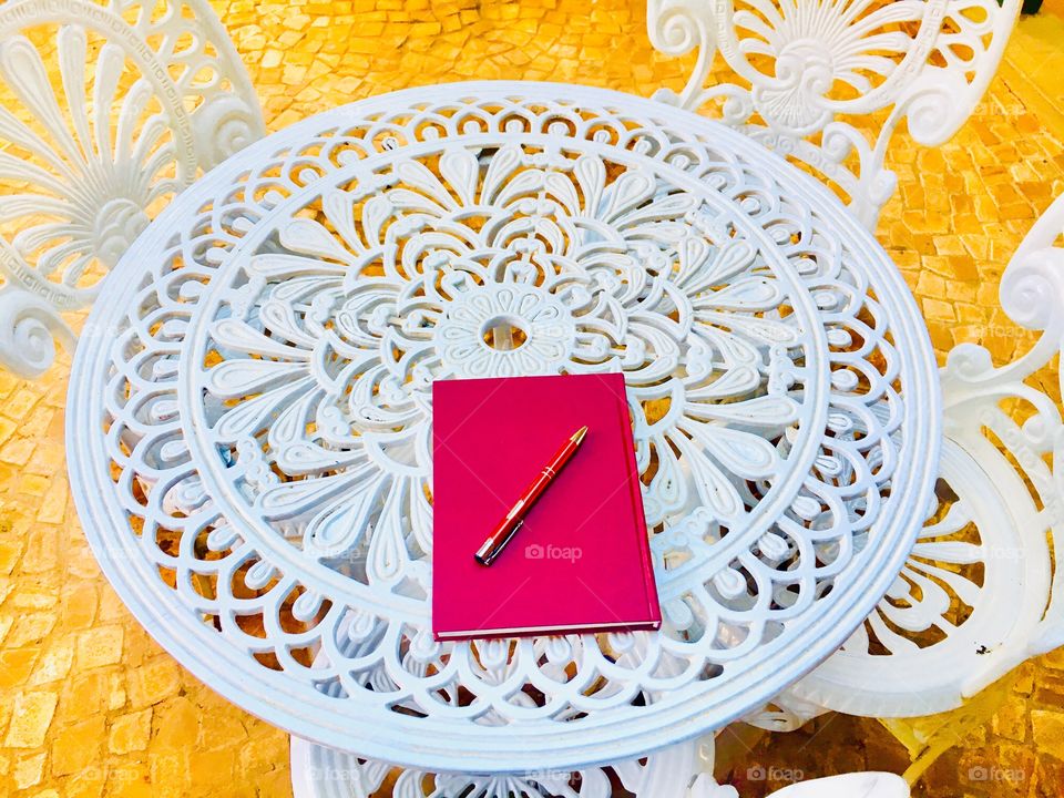 Patterns on white iron table and chairs with a notebook and pen on top of it.