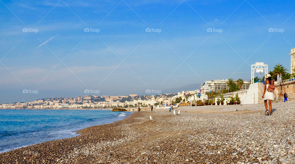 Panoramic view of bay with young black women in sundress, hat and wearing a backpack, walking on the stone beach in Nice, France
