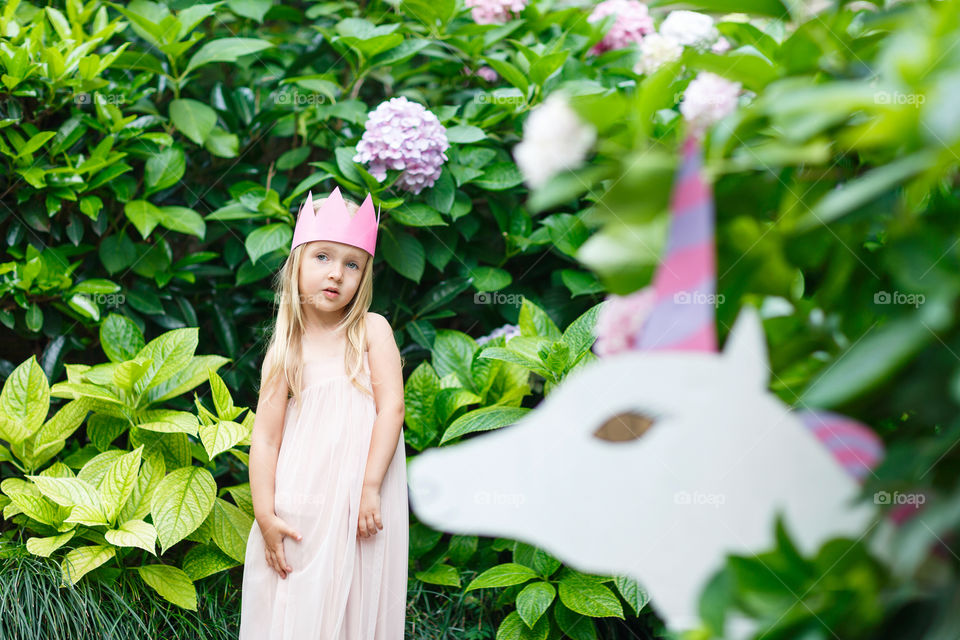 Happy little Caucasian girl with blonde hair and crown standing near unicorn in blooming garden at summer day