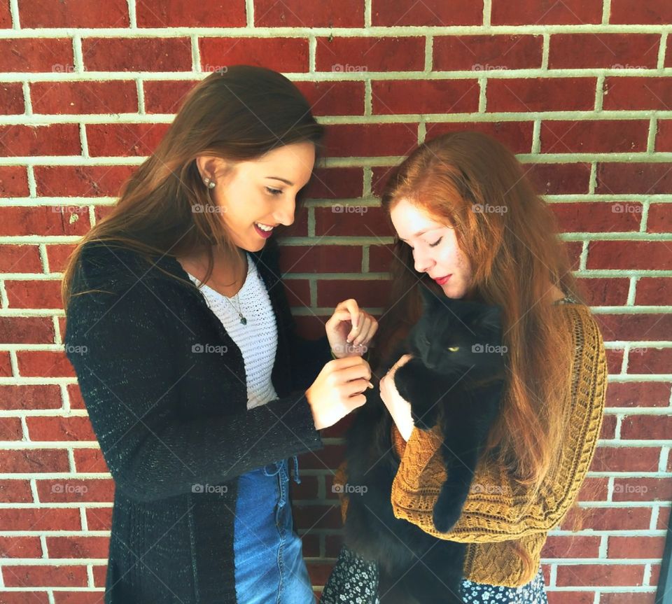 two best friends playing with a black cat in front of a brick background