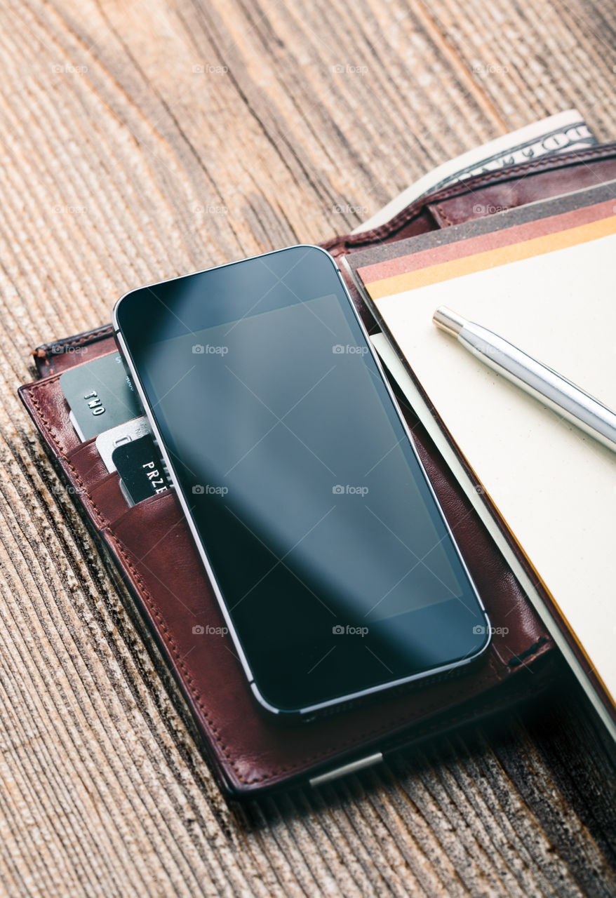 Smartphone with blank screen, wallet, dollar banknotes, debit credit cards and notebook on wooden table. View from above. Portrait orientation