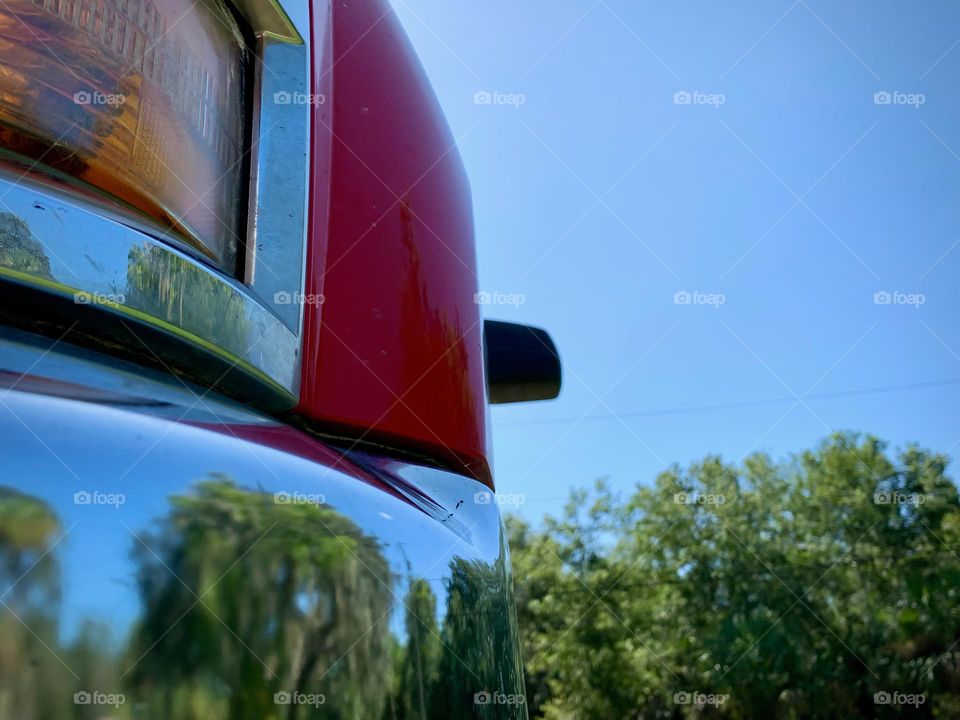 Commuting: front left side of a red pickup truck vehicle with chromed front bumper reflecting as a mirror the green trees, close up.