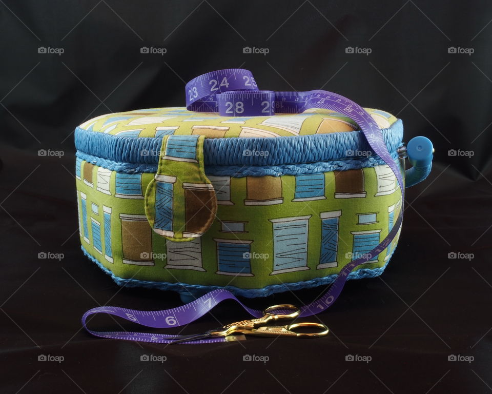 An octagon shaped sewing box with spools of thread pattern along with a purple tape measure dangling from the top and a pair of small golden handled scissors at its base.