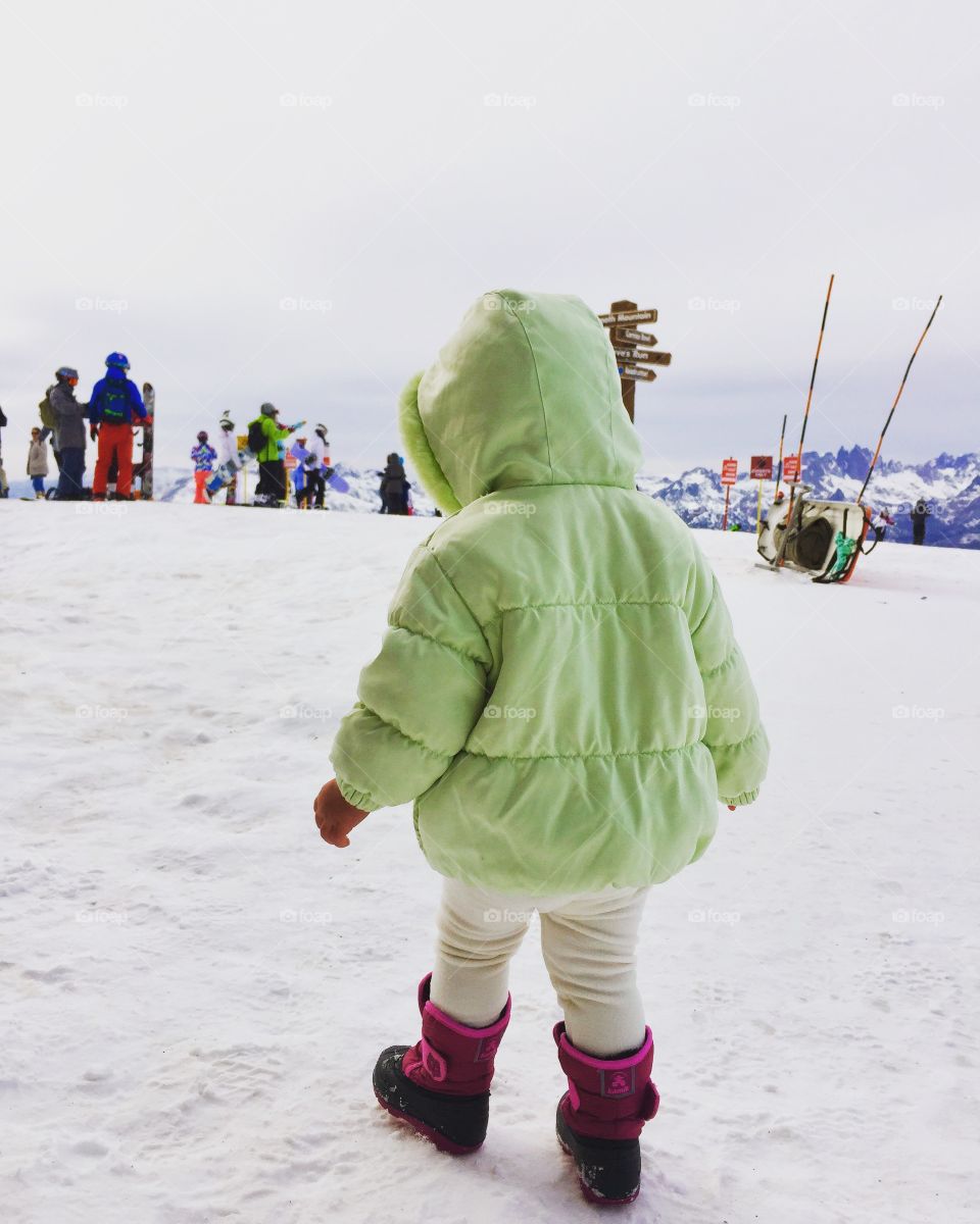 Toddler girl in green hooded winter coat with pink and purple snow boots standing on top of snow covered mountain 