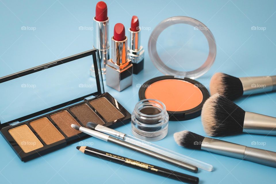 Set of cosmetics from a palette of nude eye shadow,round face powder, eye pencils, soft eyebrow tags,makeup brushes and three red lipsticks on a blue background, side view. Concept female cosmetics, beauty salon.