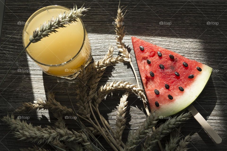 A piece of ripe, red watermelon and cold juice, a cocktail with ice and ears of wheat stand on a wooden table. Summer drinks and snacks.