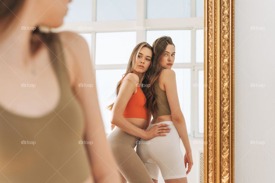 Fashion beauty models two sisters twins beautiful girls with long hair in sport wear looking to mirror in the bright studio
