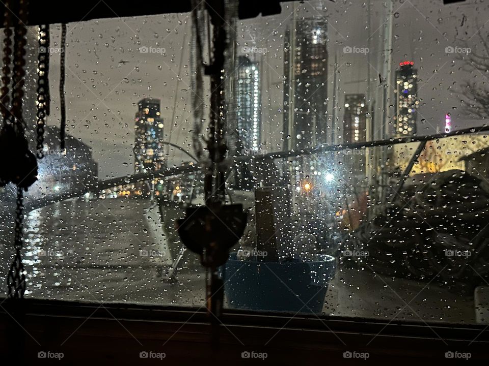 Raindrops on the window of “Salvation”, the “28 ft 1969 Luhrs” cabin cruiser we call home are illuminated by the bright lights of NYC here at “Newtown Creek” by the “Pulaski Bridge” in LIC, Queens. 2023. Hypnotic Productions