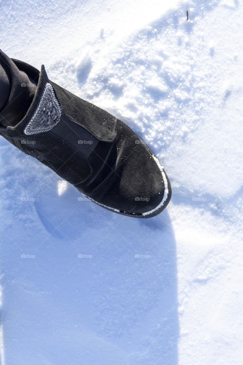 In winter, white snow lies on the street and shimmers in the sun, and a woman's foot is standing on the snow, which walks in the snow.