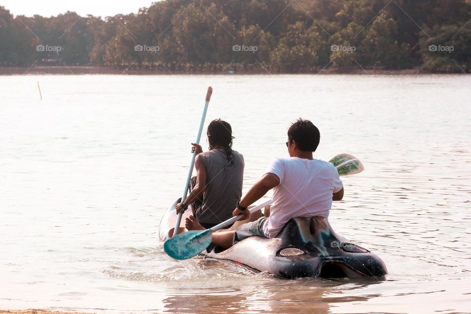 View of two adult men sitting in a canoe pedaling the paddles they are holding during the day, a backdrop of land full of trees.