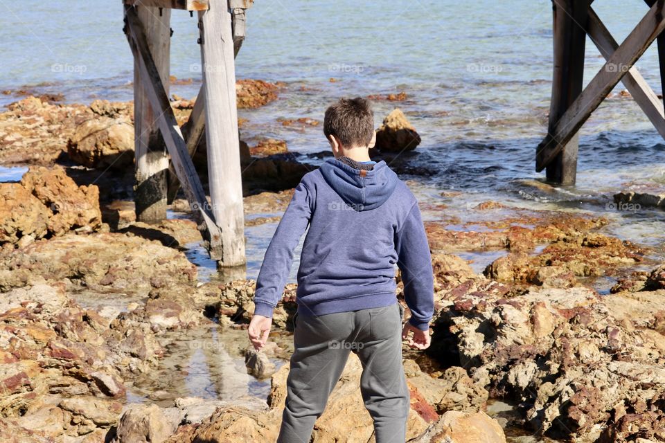 Twelve year old boy in hoodie walking across rocks near ocean Jetty wharf dock