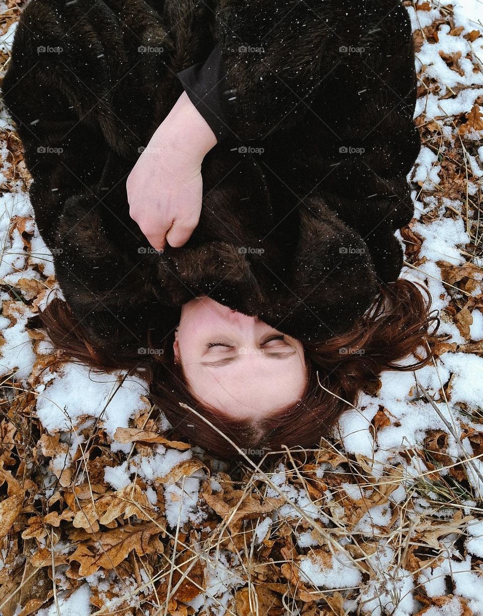 portrait of a light-skinned girl lying among the snow and autumn leaves and covering herself with a fur coat