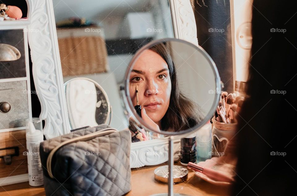 Close-up of a woman applying concealer to her face while looking into a small mirror