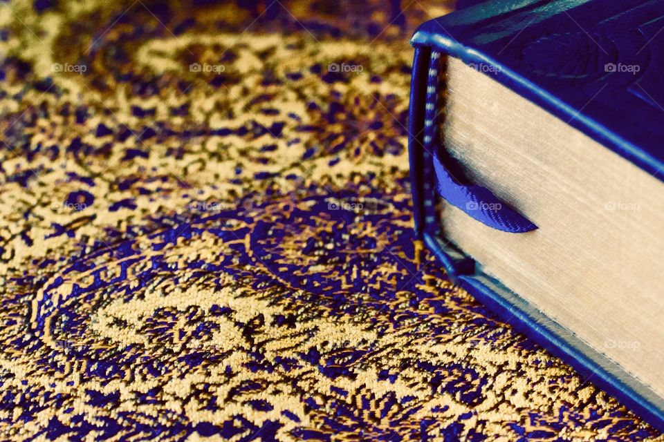 A blue with gold-leaf gilded pages and a blue ribbon bookmark on a blue and gold paisley tablecloth with interwoven shiny gold threads