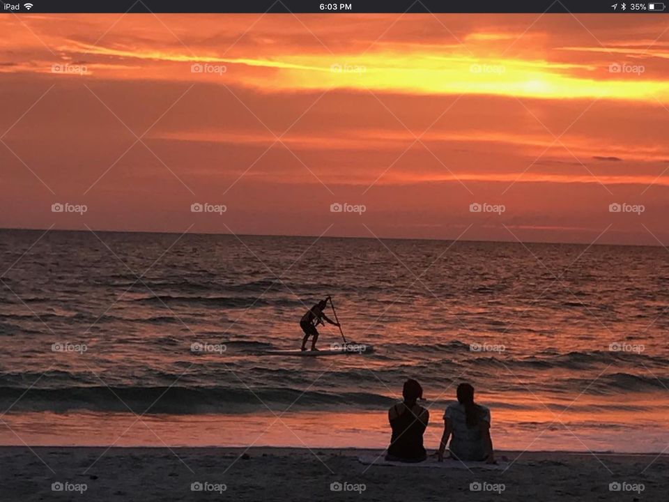 A person paddles a board along the water at sunset as a couple watches on the shore. The sky is illuminated orange with yellow streaks.