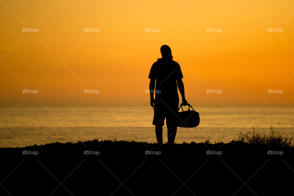 silhouette of a man holding a duffle bag at sunset by the ocean