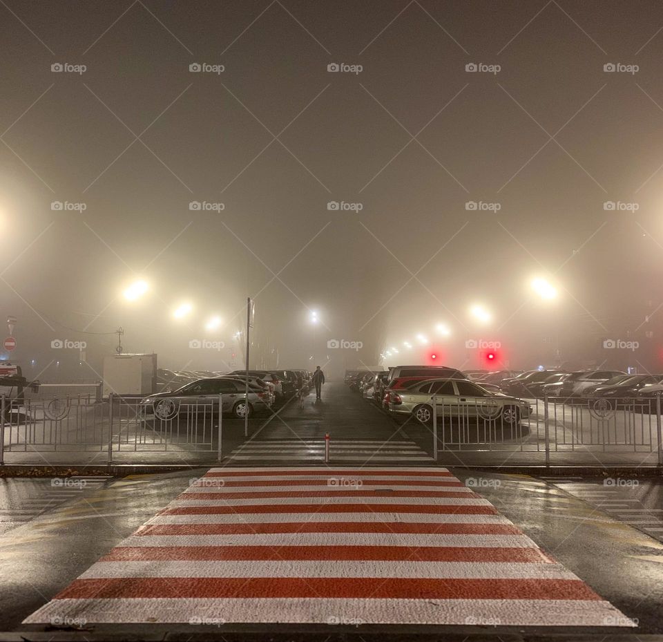 Lone figure walks alone in the foggy night through the parking lot, surrounded by soft, glowing streetlights and a misty atmosphere. Red pedestrian crossing adds contrast to the muted tones, creating a sense of solitude in the urban setting 