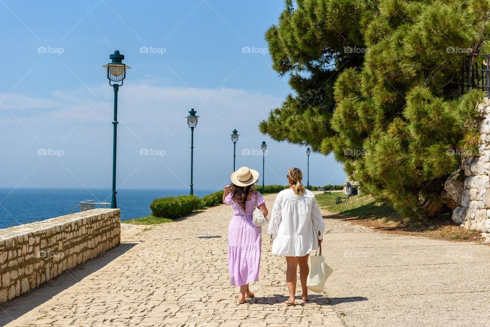 Rear view of two female friends walking on a paved path on sea coast