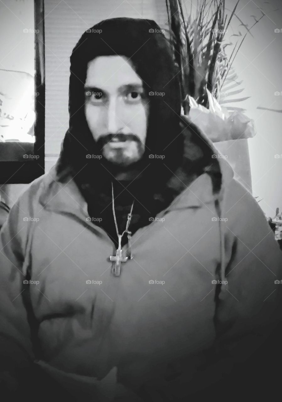 A young man with a kind, serious expression bundled up with a warm hooded jacket donning a cross pendant necklace.