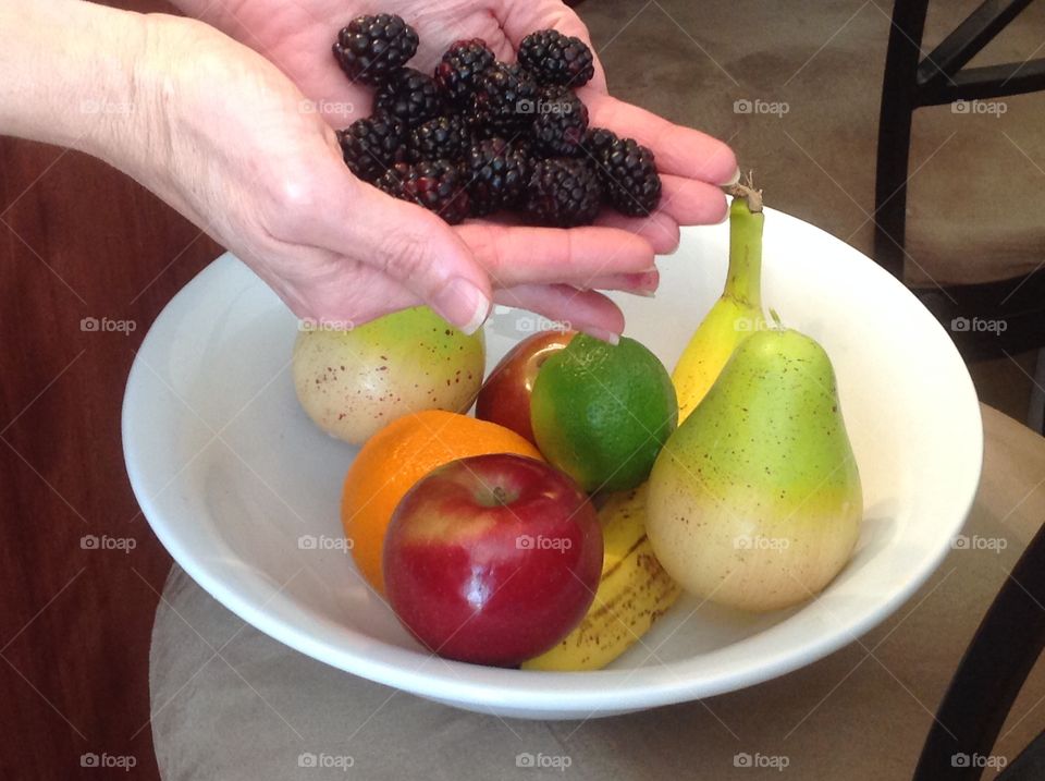 Holding in hand blackberries near a bowl of colorful fruit.