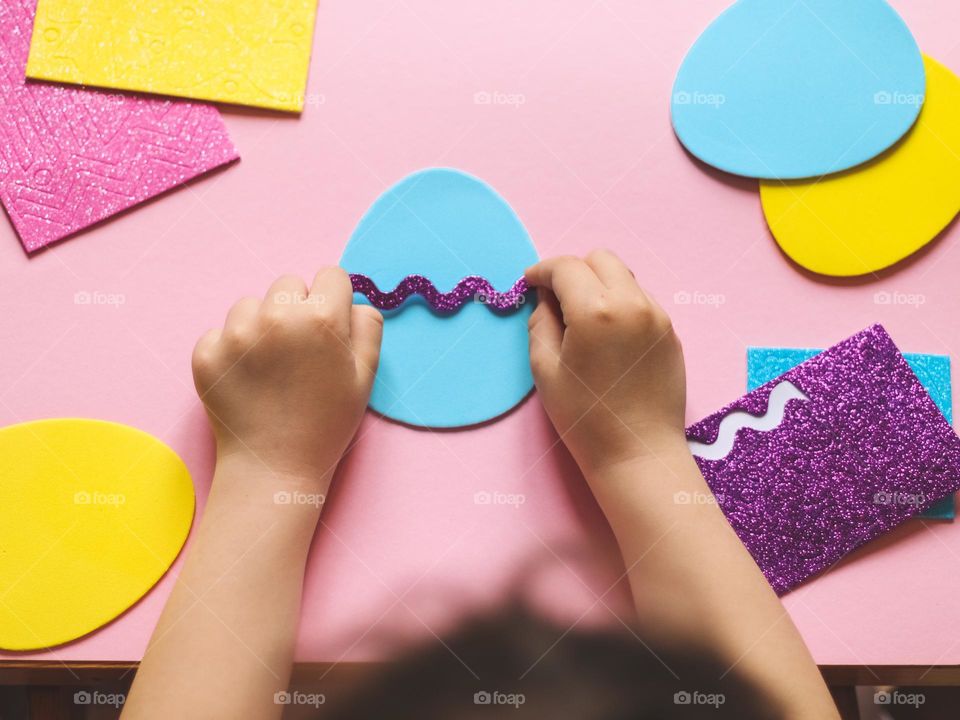 Hands of a little caucasian girl glue a lilac zigzag felt sticker on a blue egg, sitting at a pink children's table, close-up top view. Concept coative kids, easter preparation.