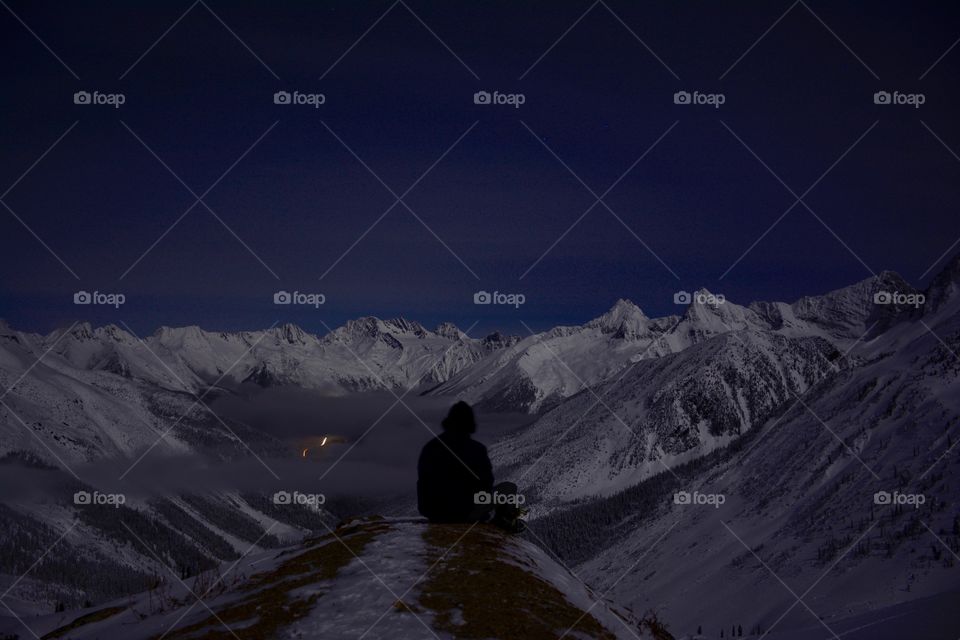 Silhouetted man sitting on rugged cliff edge atop a mountain 
