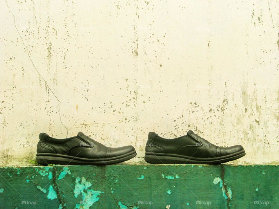 Close-up of a pair of black shoes placed on a peeling green surface, against a backdrop of a dull, cracked white wall in eye level view