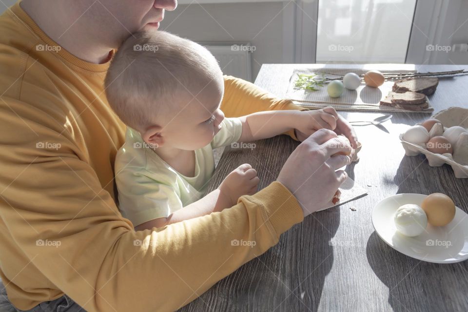 A dad and a little boy prepare for Easter at home indoors, paint Easter eggs with early-colored paints, sit at a table, sunlight falls on a wooden table.