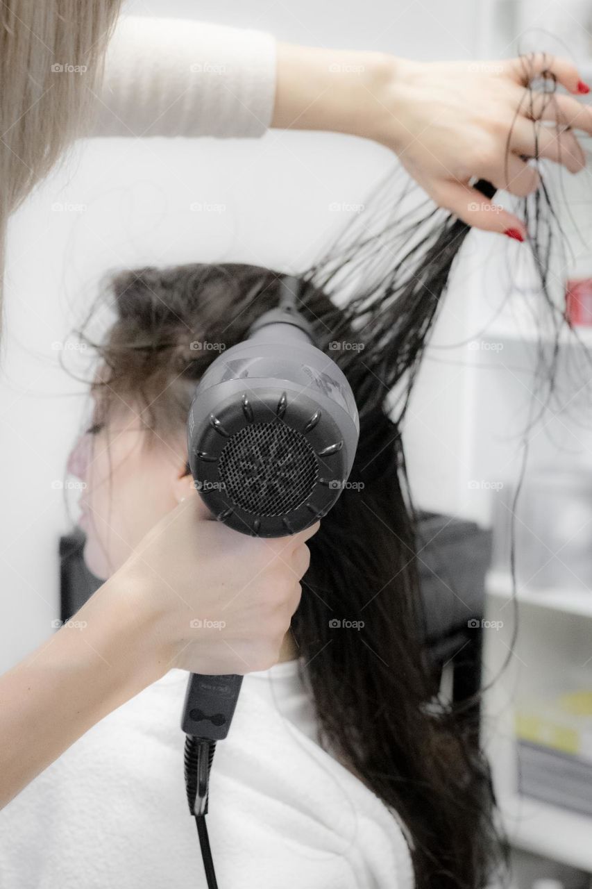 The hand of a young caucasian hairdresser girl dries with a hairdryer the long brown hair of a young client sitting in a chair with her eyes closed against the background of blurry wardrobes with hair cosmetics, side view close-up with depth of field