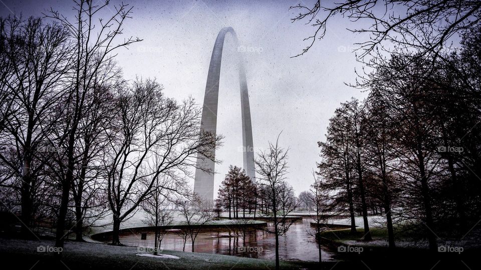 Snow falling on grounds of Saint Louis Gateway Arch National Park with lake and trees in foreground