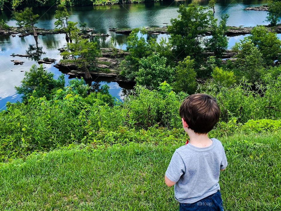 A child observes the beauty of nature , standing quietly alone as he watches the water rush over the rocks.