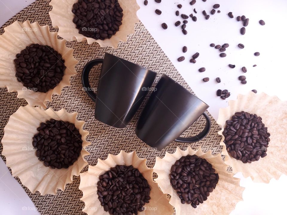 Coffee beans displayed in coffee filters surrounding two black coffee cups on beige placemat on a white surface.