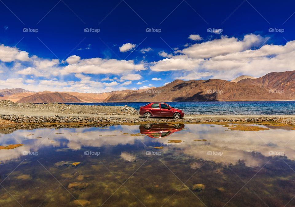 red color car parked on the road with lakes by both side of the road and cloud reflection seen on lake water
