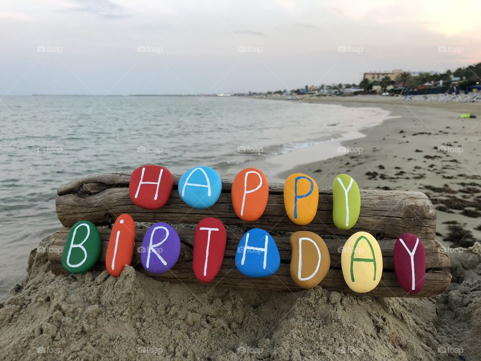 Happy Birthday message with multi colored stones over two pieces of wood with sea background