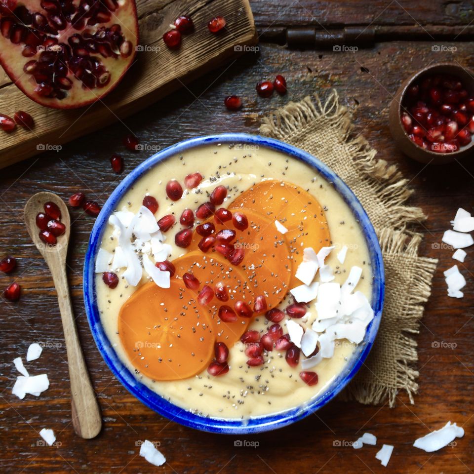 Banana smoothie bowl with sliced persimmons, pomegranate seeds and coconut flakes in a blue bowl on a rustic wood table.