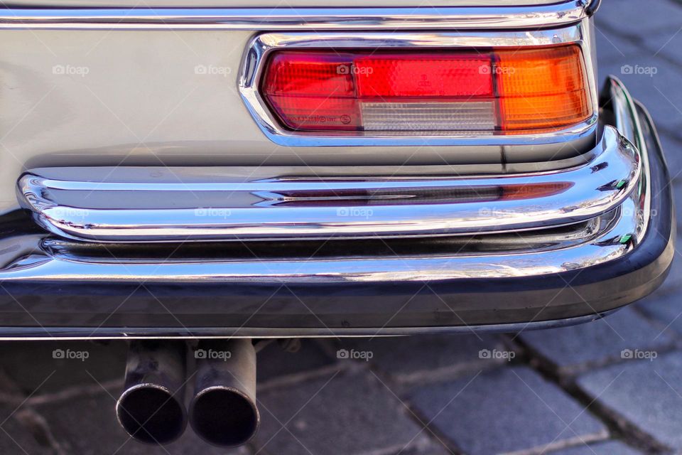 Detailed closeup of the orange taillight and a shiny silver bumper of a beige vintage car parked on cobblestones