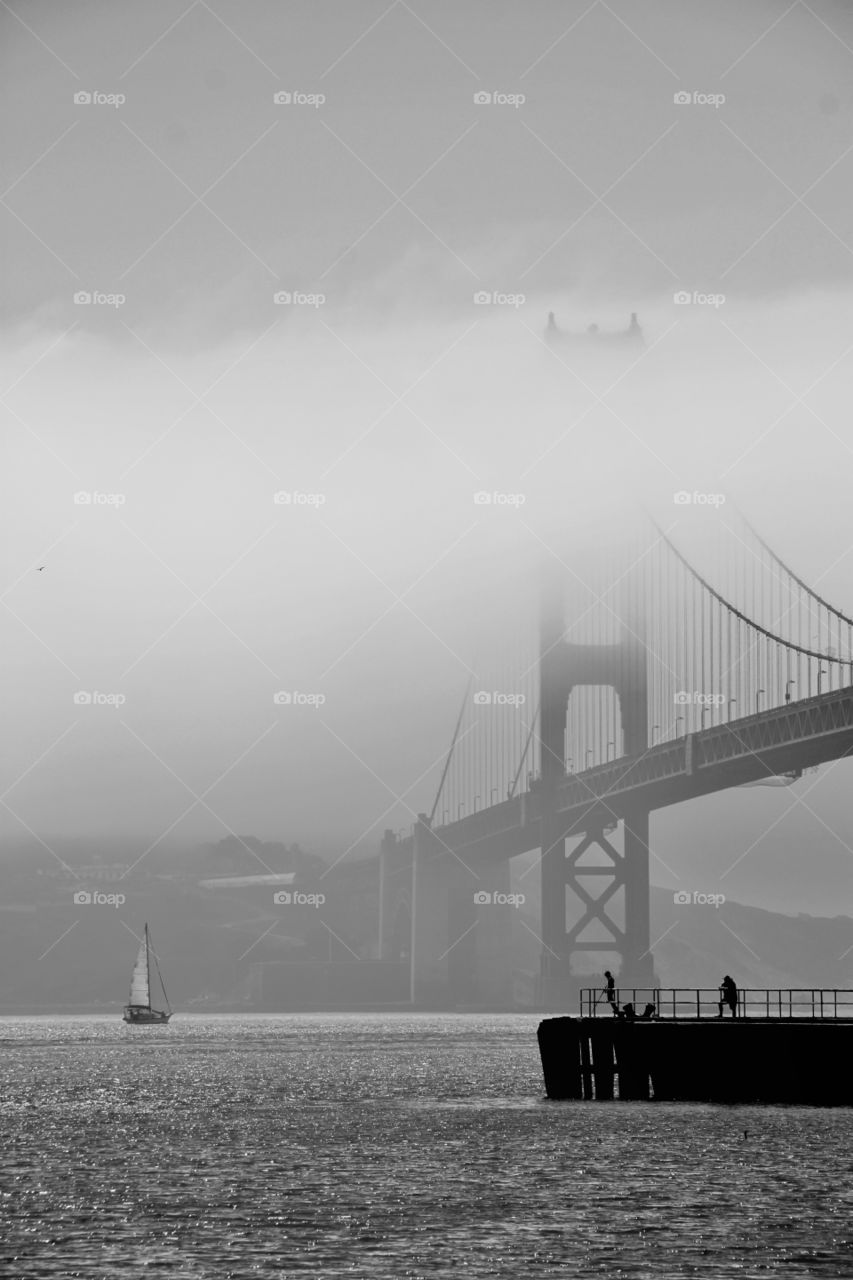 A sailing boat sliding down the sea approaching the Golden Gate Bridge, San Francisco. The mist covers the top of the bridge, giving the moment a magical touch.