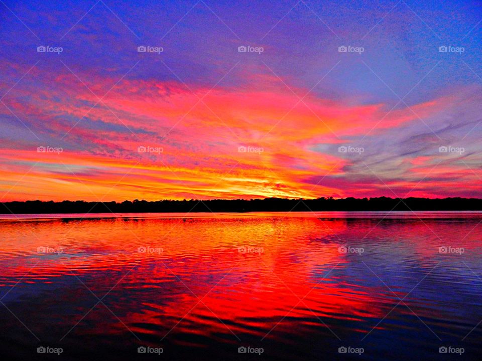 The sky features streaks of pink and orange clouds, and the colors are reflected in the wet sand and shallow water of the beach. The horizon line is visible, with the sun having just set, creating a bright glow