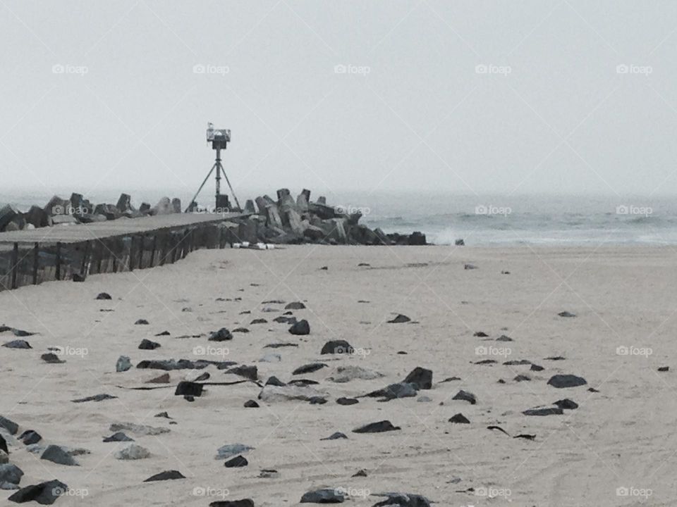 A photo of a beach in Point Pleasant Beach, NJ near the Manasquan Inlet. Rocks in the foreground make the beach resemble the moon, as does the gray sky. On the left is a pier people walk out on to fish or to just get a closer view of the ocean. 