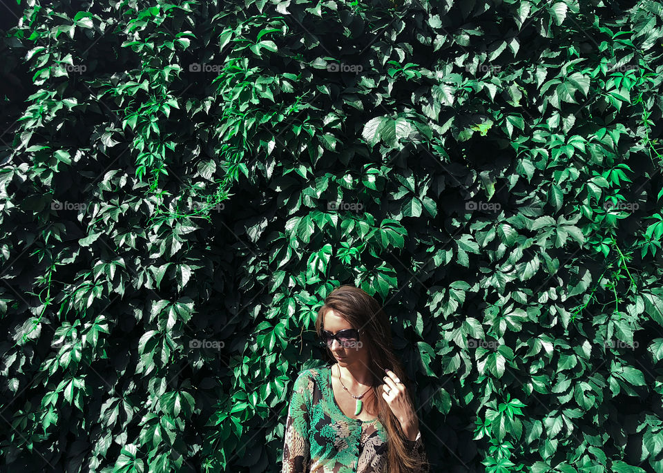 Young woman wearing green clothes standing in front of natural green wall made of green leaves 