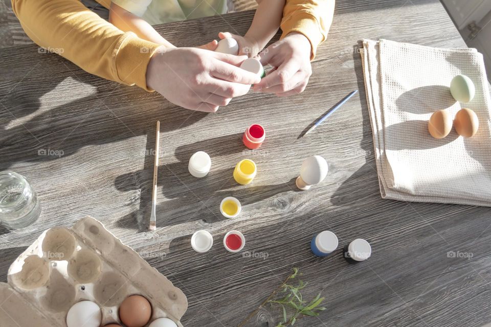 A little son and his father together in their free time paint Easter, chicken eggs with paints and brushes for the holiday. A child and a man create a craft and natural product in their family business.