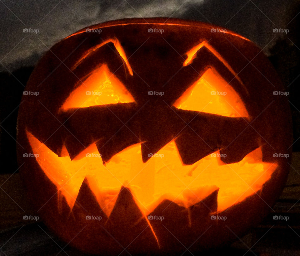 A roughly carved, lit up pumpkin with a menacing orange grin, against a dark cloudy sky