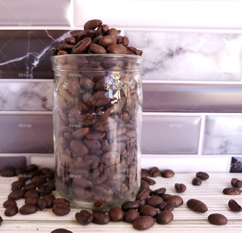 A clear glass jar with overflowing coffee beans in front of a violet gray, white and black tile backsplash.