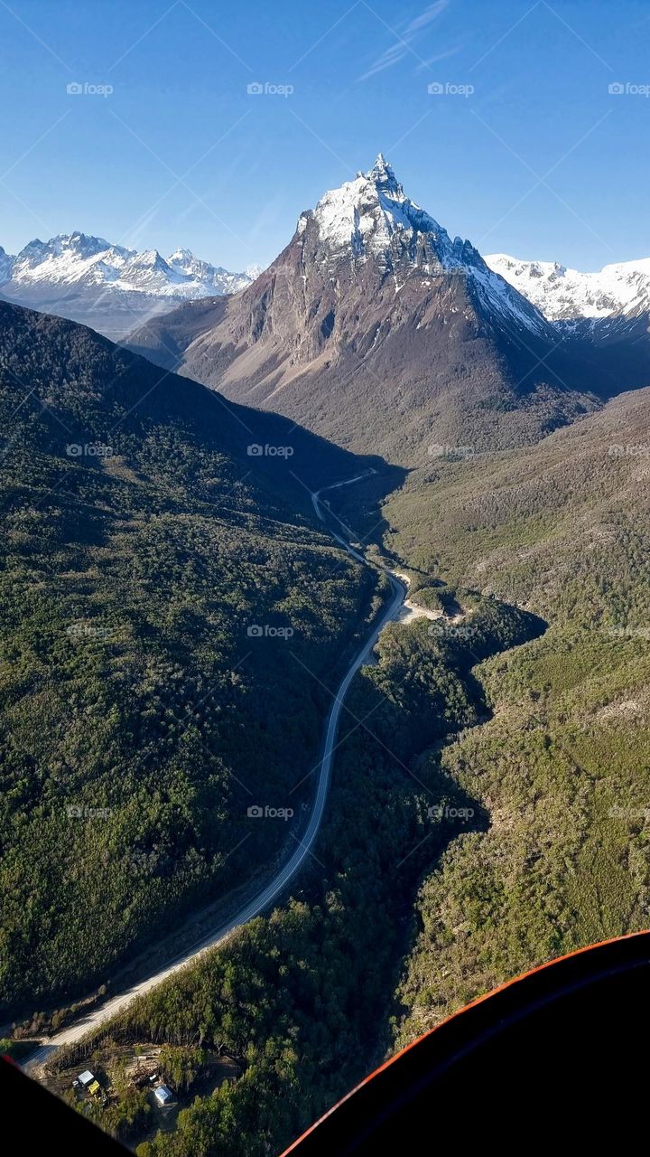 Admiring the landscape from a helicopter. In the middle, the national route through the green forest that surrounds it, snow-capped mountains and a clear sky.