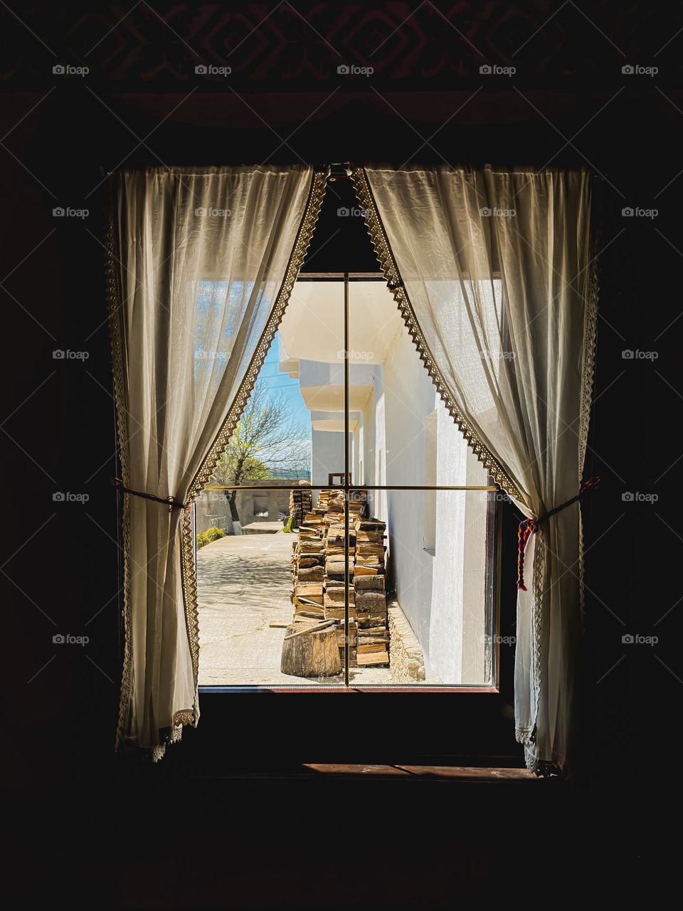 The pile of wood, chopped and laid out in the back of the house, seen from behind an old draped window, the kind you can only find in the countryside