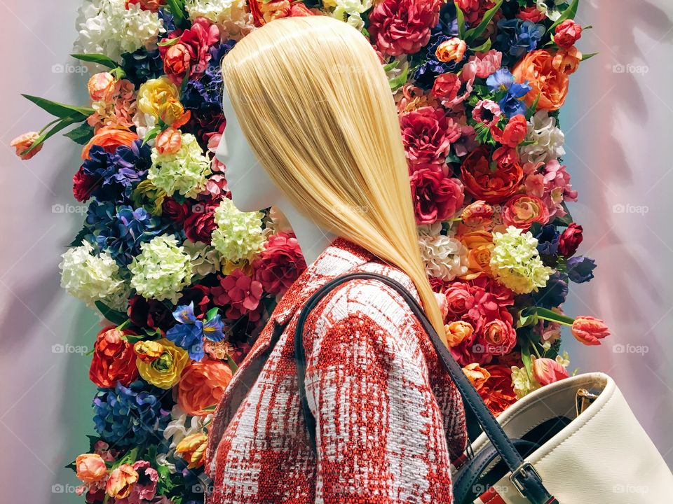 A fashionable blonde mannequin on display over a wall of plastic coloured flowers.