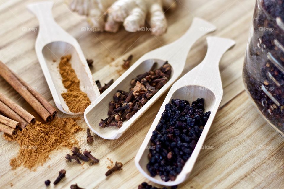 Closeup of ground cinnamon and cinnamon sticks, ginger root, whole cloves, and dried elderberries in wooden scoops on a wooden surface as ingredients for homemade elderberry syrup