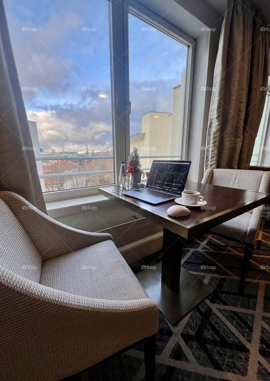 A table near the window. On the table is a laptop, coffee and water.