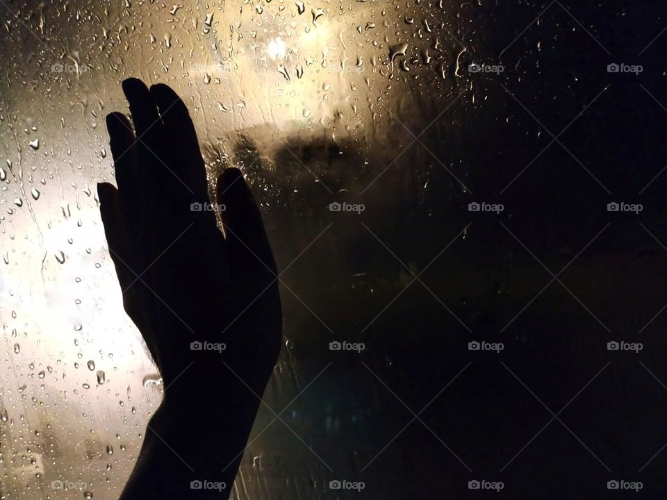 The silhouet of someone's hand watching a rainy night, iluminated by the dull glow of a lonely lamppost, through the wet glass of the window... loneliness or peace?
